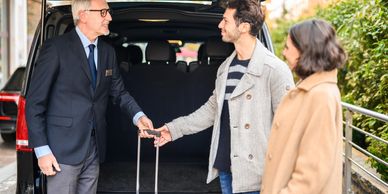 Chauffeur in a suit shaking hands with a passenger next to an open car trunk loaded with luggage